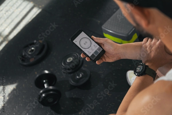 Fototapeta A man with a smart phone in the gym. Looking at the screen of the smart phone. With dumbbells on the floor. health and wellness concept.