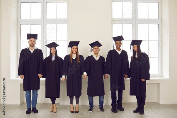 Fototapeta General portrait of a group of happy and successful multiracial university graduates in academic dresses standing holding hands. Education, graduation and people concept.