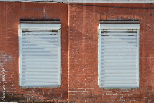 Fototapeta Shutters. Old facade painted red brick wall with 2 windows closed by white roller shutters, illuminated by bright sunlight