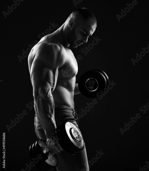 Fototapeta Muscular men bodybuilder is working out in gym, lifting alternately two dumbbells, doing exercises for biceps and looking down over black background. Young man lifting weights. Black and white 