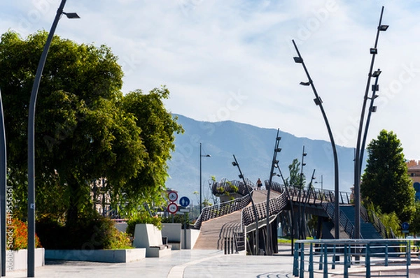 Fototapeta A city park with curved footpaths above the ground on pylons, curved lamp posts, green trees and architectural structures against the backdrop of a mountain range.