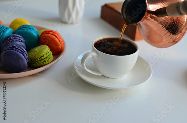 Fototapeta Pouring coffee into a cup from a turkish coffee pot, multicolored macaroons on white background