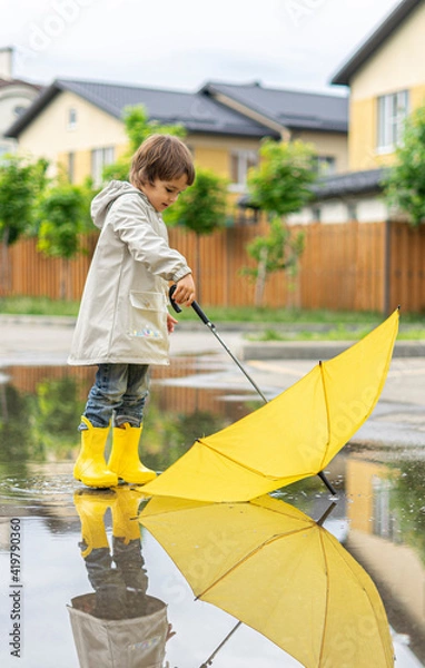 Fototapeta Boy holds yellow umbrella standing in puddle outside. On sunny day happy kid walks near the house, plays, has fun
