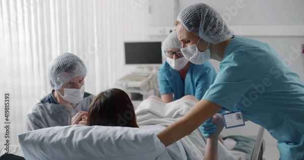 Fototapeta Woman giving birth with husband holds her hand in support and obstetricians assisting.