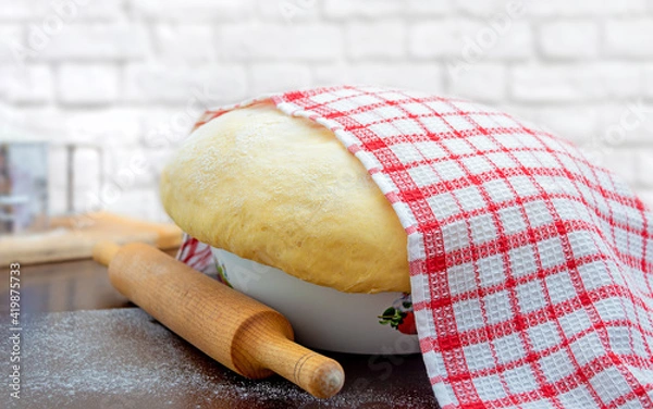 Fototapeta Raw yeast dough covered with a towel in a bowl on the floured kitchen table, recipe idea. Concept home baking or making dough.