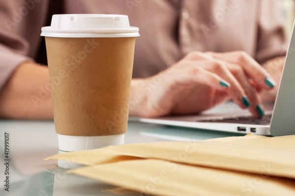 Fototapeta Young woman drinking coffee from disposable cup at the office while working with a computer. Brown paper cup with a white plastic lid 