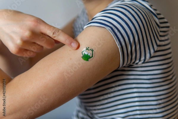 Fototapeta Woman pointing her arm with a fun, colorful, adhesive bandage after injection of vaccine or a scratch on the skin. First aid. Medical, pharmacy, and healthcare concept. After vaccination treatment.