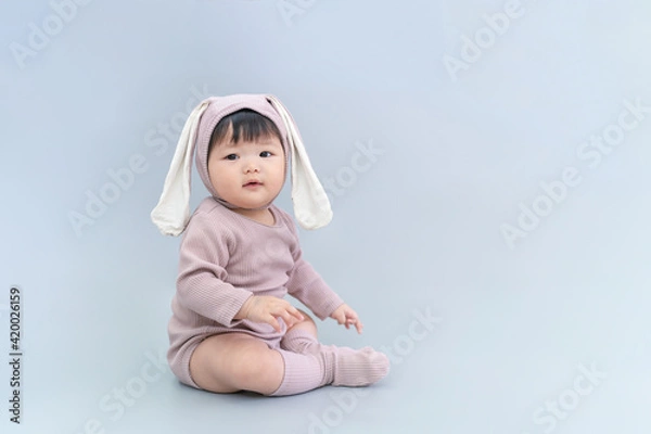 Fototapeta Easter bunny, Baby in rabbit cloth. Asian happy baby smiling, sitting on gray background. Cute 6 months baby start sitting with copy space as Easter concept, baby or kid department in hospital