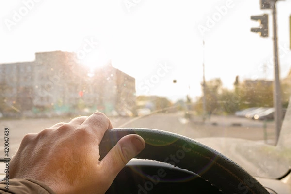 Fototapeta the driver's hand on the steering wheel of a car that is blinded by bright sunlight in the windshield while driving on the road through an intersection with a traffic light