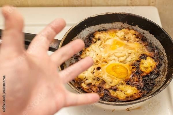 Fototapeta view of overcooked, burnt eggs, omelet on an old cast iron frying pan on an electric stove against the background of the hand of an emotionally outraged person
