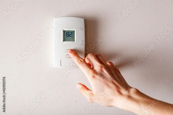 Fototapeta A woman is pressing the up button of a wall attached house thermostat with digital display showing the temperature. A concept image for electricity bill, heating, cooling, eco friendly, saving etc.