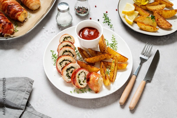 Fototapeta Bacon wrapped chicken rolls stuffed with feta cheese and spinach served with baked potato wedges and tomato sauce. Beautiful festive lunch or dinner. Gray stone background.