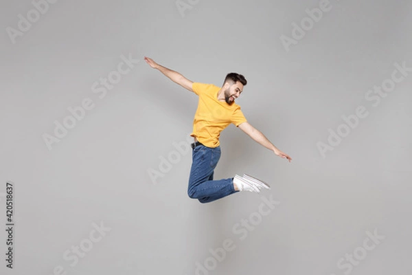 Fototapeta Full length of young bearded student smiling happy cheerful overjoyed man 20s in yellow basic t-shirt jump high looking aside flying outstretched hands isolated on grey background studio portrait