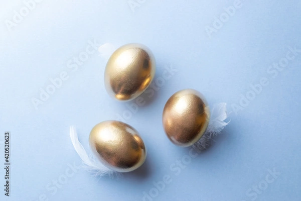 Fototapeta Golden easter colour eggs in basket with white feathers on pastel blue background in Happy Easter decoration. Spring holiday top view concept.