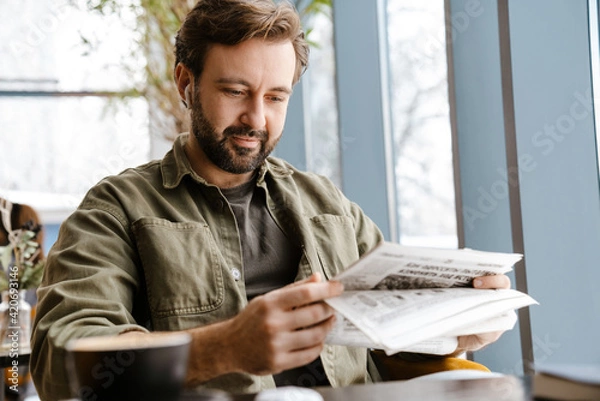 Fototapeta Unshaven focused man in earphones reading newspaper in cafe