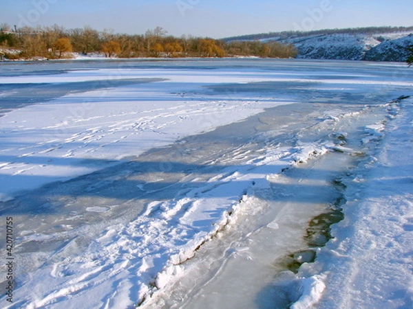 Fototapeta View of sunlit trampled trails in the snow covering the ice shell of the Dnieper shore.