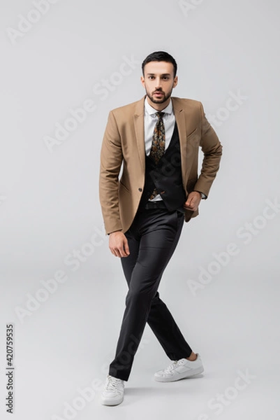 Fototapeta full length view of young muslim man in trendy suit  and sneakers looking at camera on grey