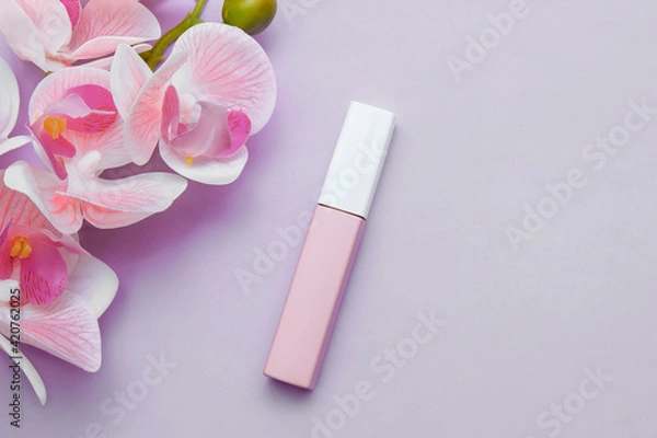 Fototapeta Closed pink lipstick on purple background with orchid flowers close-up view from the top, copy space, lip care concept, fashionable feminine and casual makeup