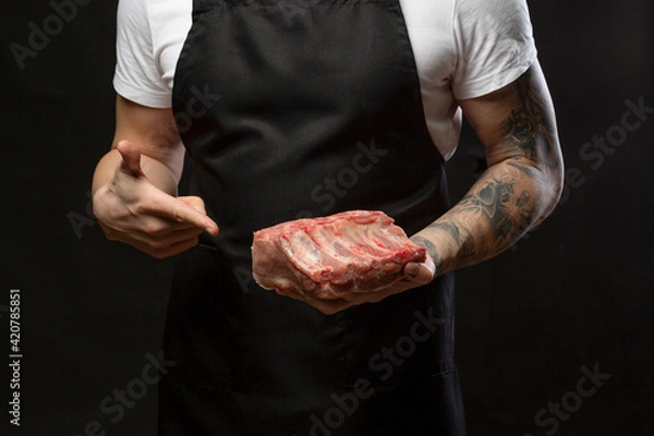 Obraz Closeup a man butcher wearing black apron pointing finger to raw pork rib. Mockup, place for text.