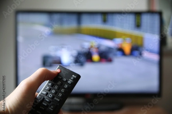 Fototapeta Watching formula 1 on TV. Thumb on the power off button on the remote (focus on the remote).