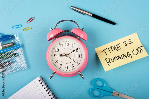 Fototapeta A pink color table clock with Time is running out words Written on a yellow sticky note and Pen with other elements on a blue background.