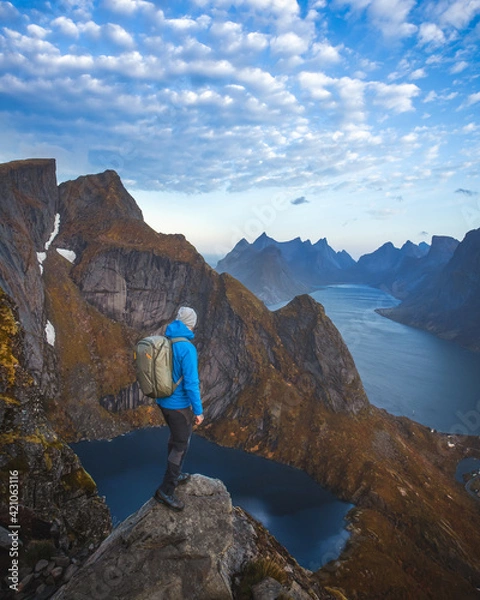 Fototapeta A man with a blue jacket, a hat, and a backpack standing on the top of the mountain, overlooking a beautiful lake and a sea in Lofoten, in Norway during the golden hour.