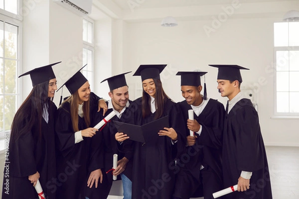 Fototapeta Group of students of different nationalities in graduation gowns look at photos in the graduation album in the university classroom. Concept of education, graduation, people, memories and emotions.