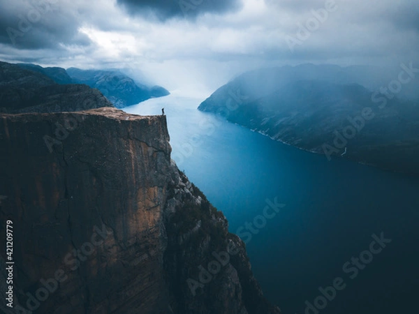 Fototapeta A man with a blue jacket, shorts, hiking boots, and backpack standing on the top of the cliff Preikestolen and overlooking a fjord in Norway. Moody landscape and sky.