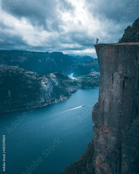 Fototapeta A man with a blue jacket, shorts, hiking boots, and backpack standing on the top of the cliff Preikestolen and overlooking a fjord in Norway. Moody landscape and sky.