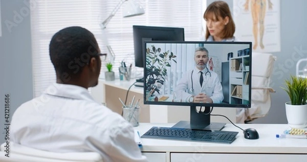 Fototapeta Close up. Over shoulder view of African American man doctor sitting in cabinet in clinic speaking with Caucasian young coworker consulting online on computer, medical web conference healthcare concept