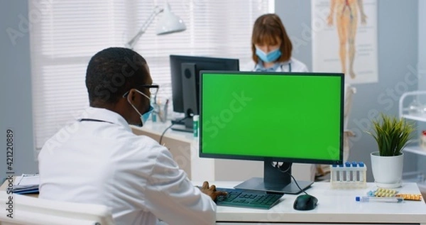 Fototapeta Back view of African American male physician sitting in hospital office and looking at green screen while working typing on computer. Medical concept, doctor browsing on monitor with chroma key