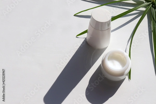 Fototapeta Face cream in glass jar, bottle of lotion and palm leaves on white table background. Hard light, shadow. Natural beauty products for skin and body care. Summer lifestyle. Top view, copy space	