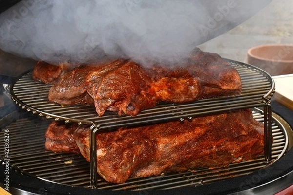 Fototapeta A large beef steak that makes smoke being cooked in the grill of an outdoor barbecue