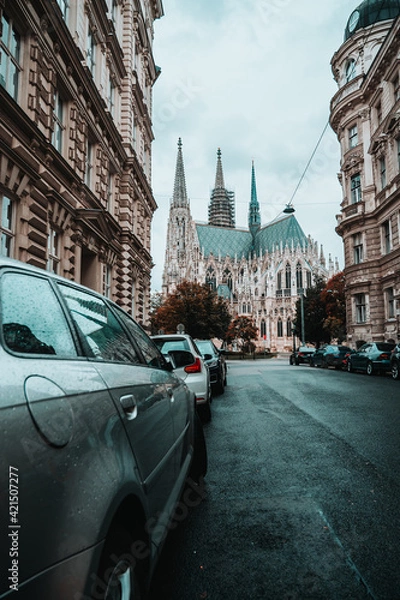 Fototapeta Beautiful streets of Austrian capital Vienna. Old gothic architecture around the narrow streets. Wet asphalt after rain and cars parked around. View on Catholic cathedral Votivkirche in downtown.