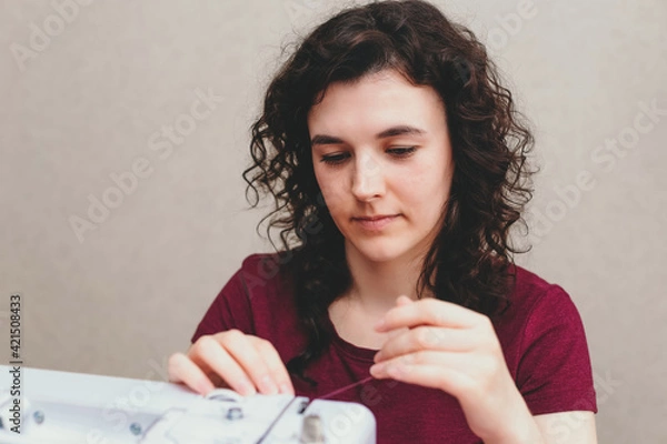 Obraz Happy attractive young woman seamstress sitting and sews on sewing machine in studio