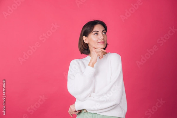 Fototapeta Beautiful woman in casual white sweater on pink red background looks to the side thoughtfully with a sweet smile isolated copy space
