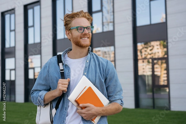 Fototapeta Smiling university student with backpack and book walking in university campus, education concept. Portrait of happy handsome man wearing stylish eyeglasses looking away on the street, copy space 