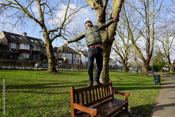 Fototapeta Portrait full body of Caucasian man  wearing green jacket, bobble hat with white pom pom walking on back of wooden bench in the park 