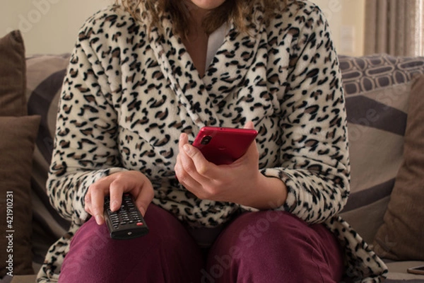 Fototapeta A young lady is sitting on a sofa with a remote tv control and a red smartphone