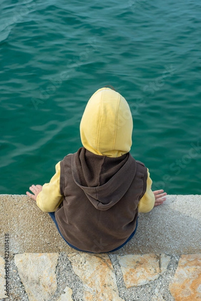 Fototapeta Kid with a yellow hat and brown vest sitting on a stone pier near the green sea water