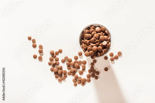 Fototapeta Chocolate callets in a white disposable cup on a white background. top view close-up. Flat lay. Tempered chocolate confectionery concept. Wholesale of chocolate.