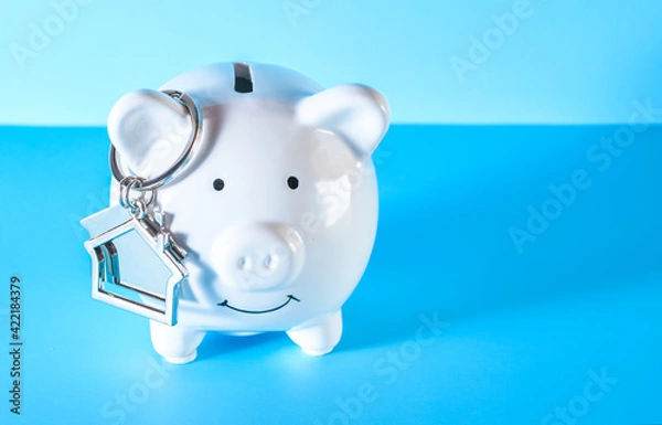 Fototapeta White piggy bank and house keychain on blue background. Saving money wealth and financial concept