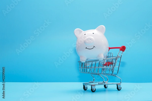 Fototapeta White piggy bank and shopping cart on blue background . Saving money wealth and financial concept