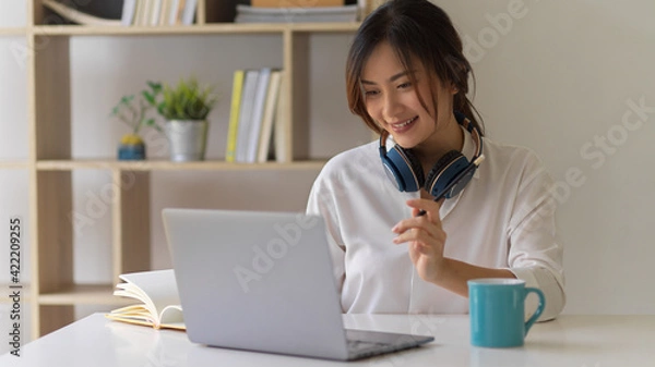 Fototapeta Smiling female student doing assignment with laptop and books in living room