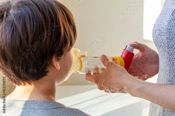 Fototapeta A caucasian woman is giving emergency asthma medication to a small kit during an attack. She uses aerosol chamber with valve mask to deliver bronchodilators or corticosteroids to relieve symptoms.