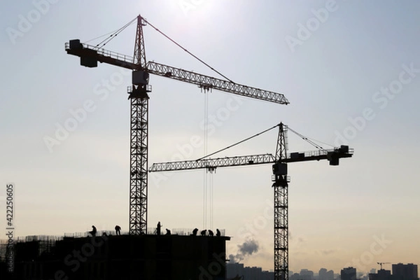 Fototapeta Silhouettes of construction cranes and workers on unfinished residential building against sunshine. Housing construction, apartment block in city