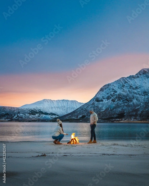 Fototapeta A couple enjoying the fire at the beach in Norway during the Blue hour,  enjoying a sunset with a beautiful colourful sky and majestic mountains behind them.