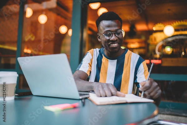 Fototapeta Happy dark skinned male copywriter writing text ideas in notebook working remotely in street cafe, successful hipster blogger in optical eyeglasses making education notes during laptop e learning