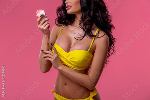 Obraz A beautiful sexy brunette in a yellow bikini holds melted ice cream in her hand. The concept of summer, warmth and good mood. Woman's portrait close-up on a background