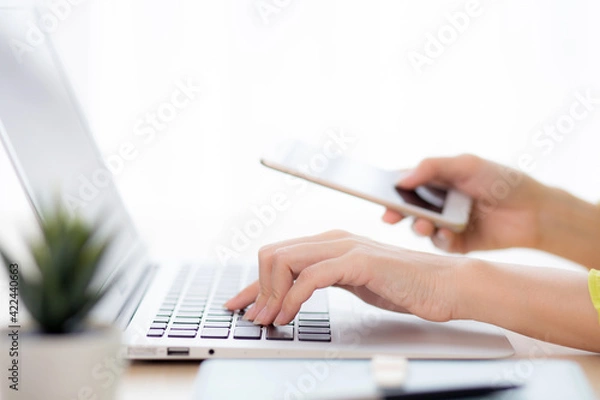 Fototapeta Closeup hand of young asian businesswoman working on laptop computer on desk at home office, freelance woman looking message on smart phone and typing on notebook, business and communication concept.